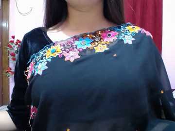 A video of a woman wearing a black saree with colorful floral embroidery, partially covering her shoulders, in a well-lit room with red curtains and floral decorations.