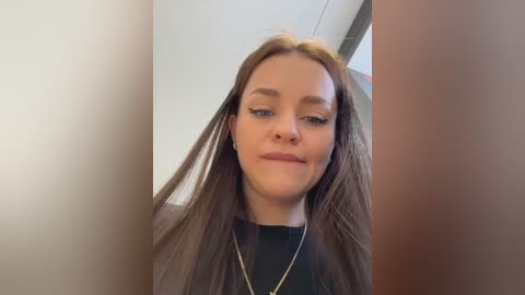 Video of a young Caucasian woman with long brown hair, wearing a black top and gold necklace, smiling slightly, indoors with blurred background.