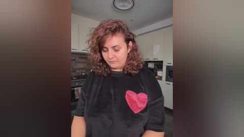 Video of a curly-haired woman with a medium build in a modern kitchen, wearing a black t-shirt with a large pink heart graphic, looking down with a contemplative expression.