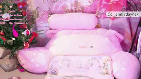 Media: Video of a pink, plush, child-sized bed with a teddy bear and pillow, adorned with Christmas ornaments, in a room with pink floral wallpaper.