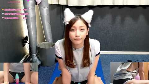 Video of an Asian woman in a white t-shirt and cat ears, sitting on a blue mat, surrounded by text chat bubbles, in a room with a gray wall and white curtains.