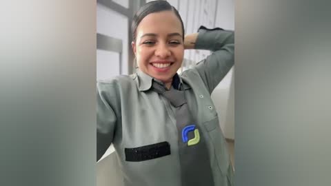 Video of a smiling Latina woman with medium skin tone and dark hair, wearing a grey work uniform with a logo on the chest, standing in a modern office.