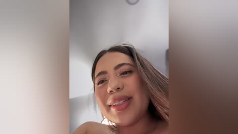 Video of a smiling, light-skinned Latina woman with long, straight hair and dark eyebrows, wearing minimal makeup. The background is a soft-focus white room, giving a warm, intimate atmosphere.