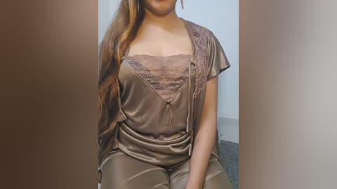 Video of a woman with long, light brown hair, wearing a satin, taupe-colored robe with lace trim and a matching camisole, seated on a grey carpeted floor.