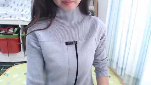 Video of a light-skinned woman with long dark hair, wearing a grey ribbed turtleneck sweater with a black zipper. Background includes a white countertop with a red bin, blue curtains, and a green rug.