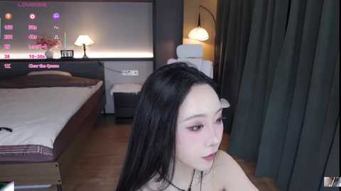 Media: A video of an Asian woman with long black hair, wearing pink lipstick, sitting on a bed in a dimly lit bedroom with a TV showing a pink screen and a dark green curtain.