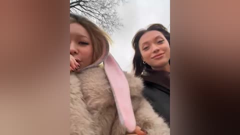 Media: Video of a young girl with a pink bunny ear headband and a woman with short brown hair, both smiling outdoors against a blurred, wintry background.