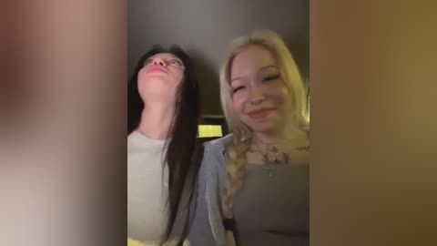 Video of two women standing side by side, smiling. One has long black hair, the other has blonde hair. They're wearing casual outfits, standing indoors with blurred background.