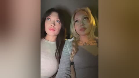Video of two young women with fair skin and long hair, one brunette, one blonde, wearing light-colored tops, looking contemplative in a dimly lit room.