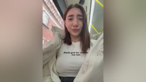Video of a young woman with straight, shoulder-length brown hair, fair skin, wearing a white shirt with \"thank god for mom me hot\" printed on it, and a beige jacket, standing in a subway car.
