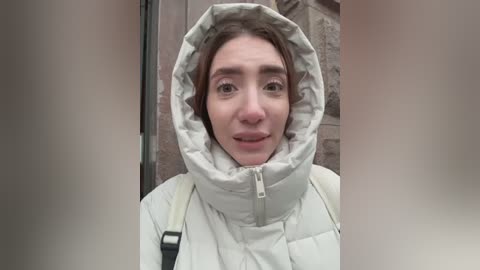 A video of a young woman with light skin, brown hair, and a neutral expression, wearing a puffy white winter coat with a hood. Background features a stone wall and a doorway.