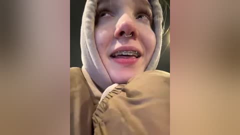 Video of a young woman with pale skin, wearing a beige hoodie and a nose ring, smiling with braces, set against a blurred background.