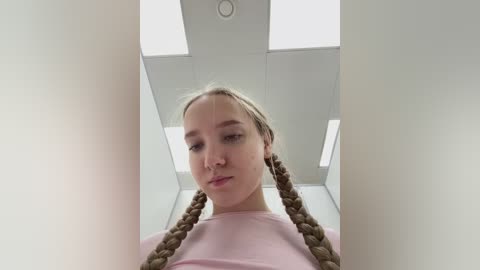 Video of a young Caucasian woman with fair skin, light brown hair braided into two pigtails, wearing a pink shirt, standing in a brightly lit, minimalist room with white walls and square ceiling lights.