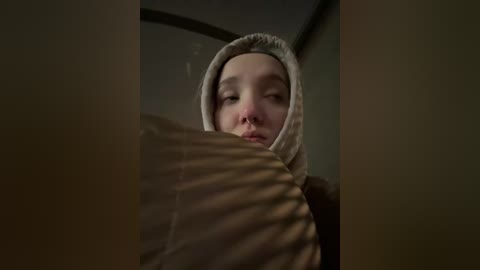 A video featuring a young boy with fair skin and a serious expression, partially obscured by a textured, beige hood. The background is dimly lit, with shadows from window blinds casting striped patterns on the scene.