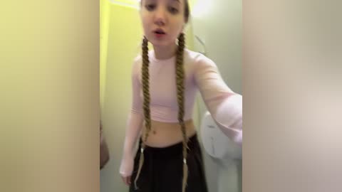 Video of a young woman with long, braided hair, wearing a pastel pink crop top and dark pants, standing in a narrow, yellow-lit hallway.