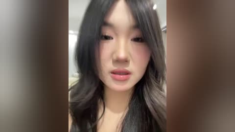 Video of an East Asian woman with long, wavy black hair and fair skin, wearing subtle makeup. Background is blurred, focusing on her face.