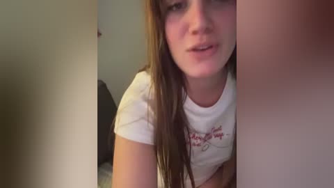 A video of a young woman with long brown hair, wearing a white T-shirt with red text, smiling, in a dimly lit room.