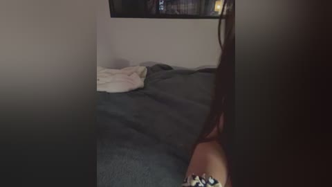 A dimly lit video of a messy, unmade bed with gray sheets and a white pillow. A figure with long brown hair is partially visible in the foreground, and a small, dark object lies on the floor.