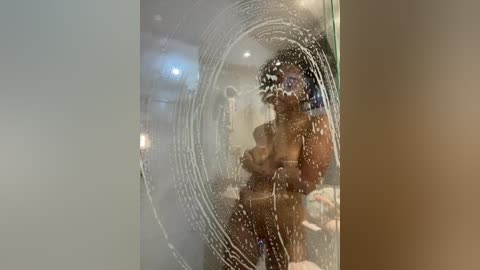 A video shows a nude person with a medium build, light skin, and dark hair, reflected in a foggy glass shower door. Water droplets form concentric circles. The background includes a white towel and a shower head.