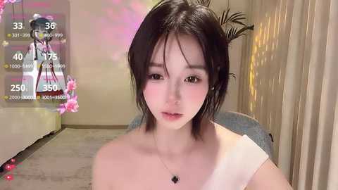 A video of an East Asian woman with fair skin, shoulder-length dark hair, and brown eyes, wearing a beige off-shoulder top. Background includes a monitor displaying game statistics, a flower arrangement, and a potted plant.