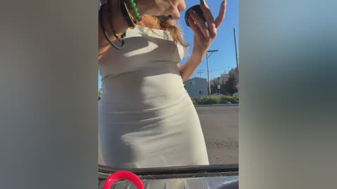 Video of a woman in a white dress taking a selfie outdoors, wearing colorful bracelets, against a suburban street background.