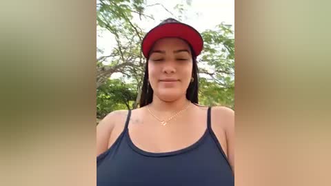 A video of a young woman with medium brown skin and long dark hair, wearing a dark blue spaghetti-strap tank top, red visor, and gold necklace, standing outdoors with lush green foliage in the background.