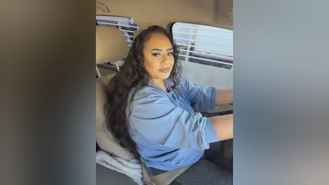 Video of a light-skinned woman with long, wavy black hair, wearing a light blue shirt, driving a car. She has a neutral expression, and the car's interior is visible.
