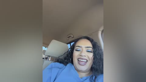 Video of a happy, middle-aged woman with long, curly black hair and medium brown skin, wearing blue eyeshadow, smiling widely inside a car.