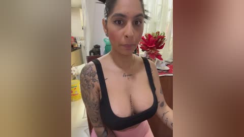 Video of a Latina woman with dark hair tied back, wearing a black tank top that reveals cleavage, tattoos on her arms and chest, sitting in a cluttered kitchen with red flowers and a yellow bucket visible.