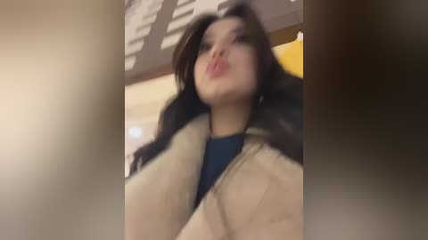 A blurry video of a woman with long dark hair, wearing a beige fur coat and a blue shirt, making a kissy face.