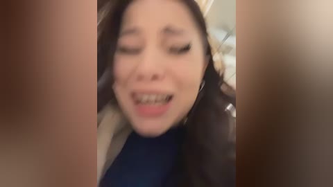 A blurred video shows an Asian woman with long dark hair, closed eyes, and a distressed expression, possibly in pain. She wears a black top and sits in a dark room with indistinct furniture in the background.