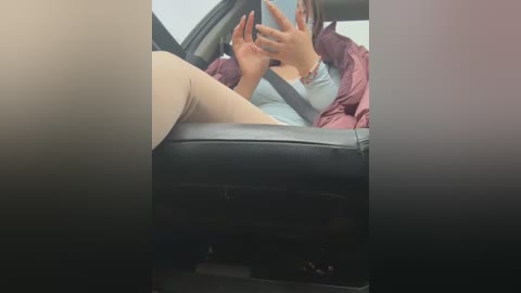 Video of a young woman with medium brown skin, wearing a pink puffer jacket and beige pants, sitting in a car, holding a phone to take a selfie, partially obscured by a car seat.