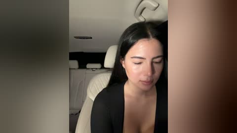 A video of a young woman with long black hair and fair skin, wearing a black top, leaning against a white leather chair in a dimly lit room.