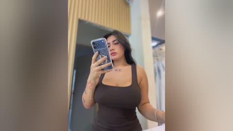Video of a curvy woman with long black hair, large breasts, and tattoos, taking a mirror selfie in a modern bathroom. She wears a tight black tank top and has a confident expression.