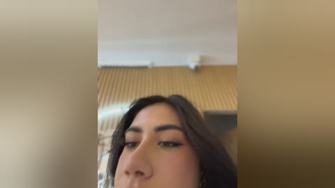 A close-up video of a young woman with medium brown skin, long dark hair, and a slender face, partially blurred. She has a neutral expression and is biting her lower lip. Background features a beige wall with a white ceiling fan.