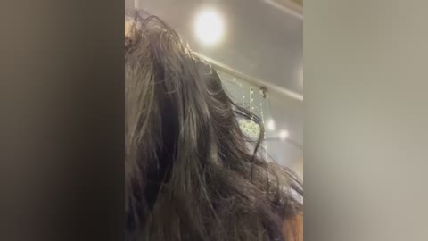 Video of a person's shoulder-length, wet, dark brown hair, viewed from the side, partially obscured by a towel. The background features a blurred, indoor setting with overhead lighting.