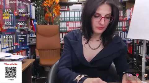 Video of a woman with long black hair, wearing glasses and a black blazer, seated at a desk in a library, with bookshelves and a vase of flowers in the background.
