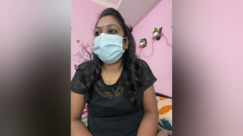 Video of a woman with medium brown skin, long black hair, wearing a black lace top, blue surgical mask, and sitting in a pink room with floral decor, plants, and a colorful blanket.