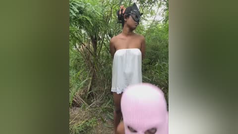 Video of a slender, dark-skinned woman with a medium build wearing a white, strapless top and black headgear, standing in lush greenery.