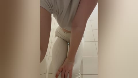 Video of a woman in white pants and a grey shirt, bending over in a tiled bathroom, with her legs partially obscuring the view.