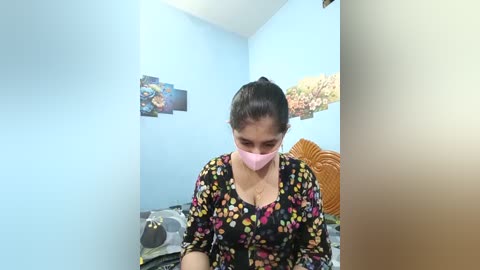 Video of a woman with dark hair, wearing a pink face mask, black floral blouse, and sitting on a bed with a grey duvet. The room has light blue walls, a wooden headboard, and colorful art.