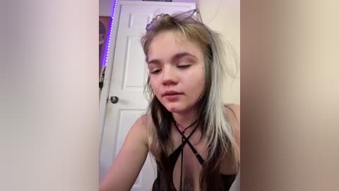 Video of a young woman with light skin and dyed blonde hair, wearing a black, strappy lingerie top, standing indoors with a white door and purple LED strip lighting in the background.