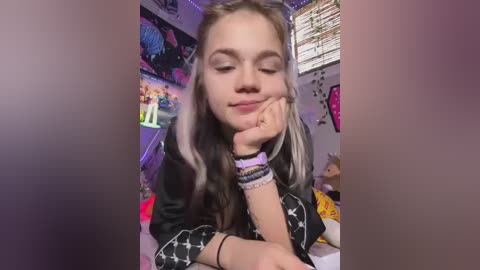Video of a young woman with long, dark hair and light streaks, wearing a black shirt with white stars, resting her chin on her hand, in a colorful, cluttered room with anime posters and toys.