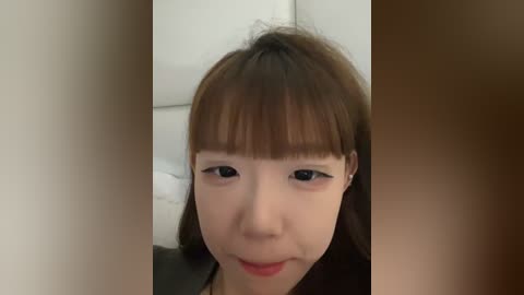 A video of a young Asian woman with straight, brown hair and bangs, wearing light makeup and a neutral expression. She has a light skin tone.