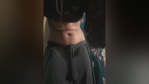 A dimly-lit video of a woman's midriff, wearing dark pants, showing a flat stomach and navel. Background is indistinct but suggests a cozy, possibly indoor setting.