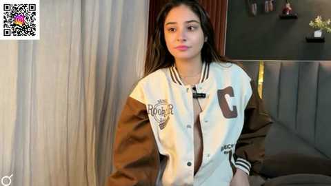Video of a young woman with long, dark hair, wearing a brown and white varsity jacket with \"C\" and \"P\" logos, in a modern, dimly lit bedroom.