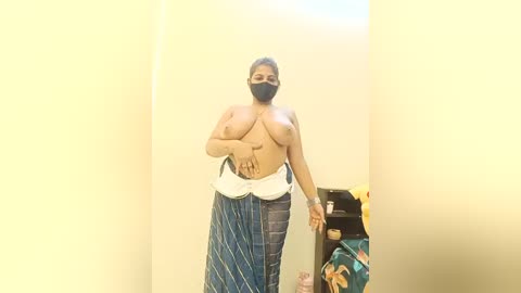 A video of a topless, dark-skinned woman with medium breasts, wearing a blue and white striped skirt, black face mask, and black hair, standing in a dimly lit, beige-walled room with a black shelf in the background.