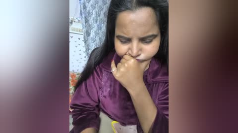 Video of a woman with long black hair, wearing a purple satin shirt, covering her mouth with her right hand, in a dimly lit room with floral wallpaper.