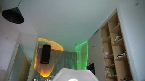 A video of a modern, minimalist bedroom with white walls, wooden built-in shelves, a black ceiling lamp, and a green ambient light highlighting the wall and bed.