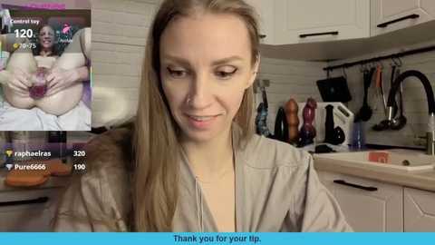 Video of a smiling, light-skinned woman with long blonde hair in a beige robe, standing in a modern kitchen with white cabinets and utensils.